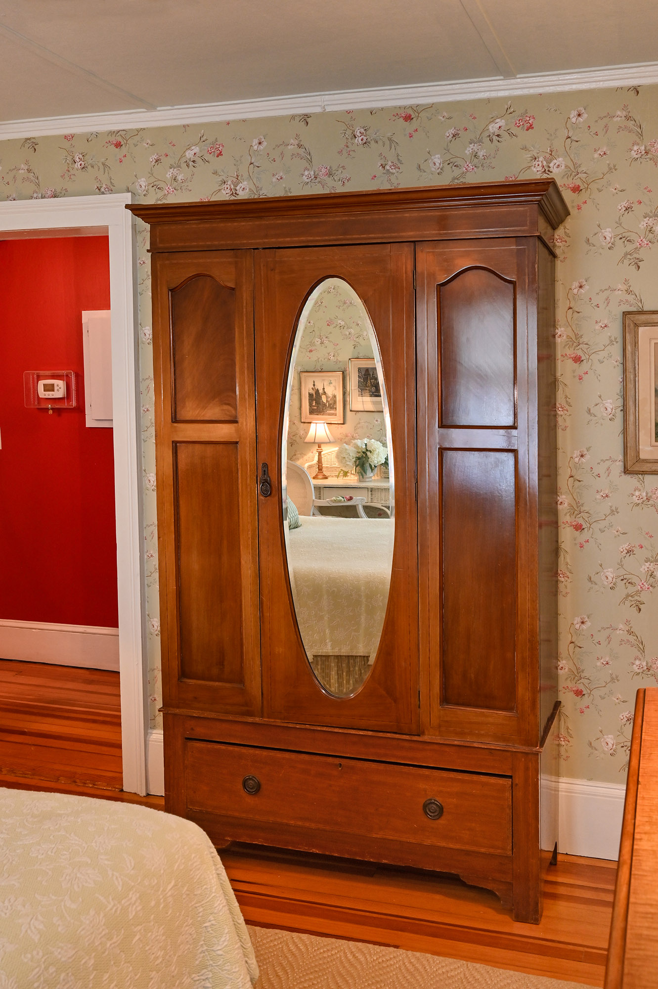 Wood wardrobe with long oval mirror on door next to open exit door. Red walls in the hallway.