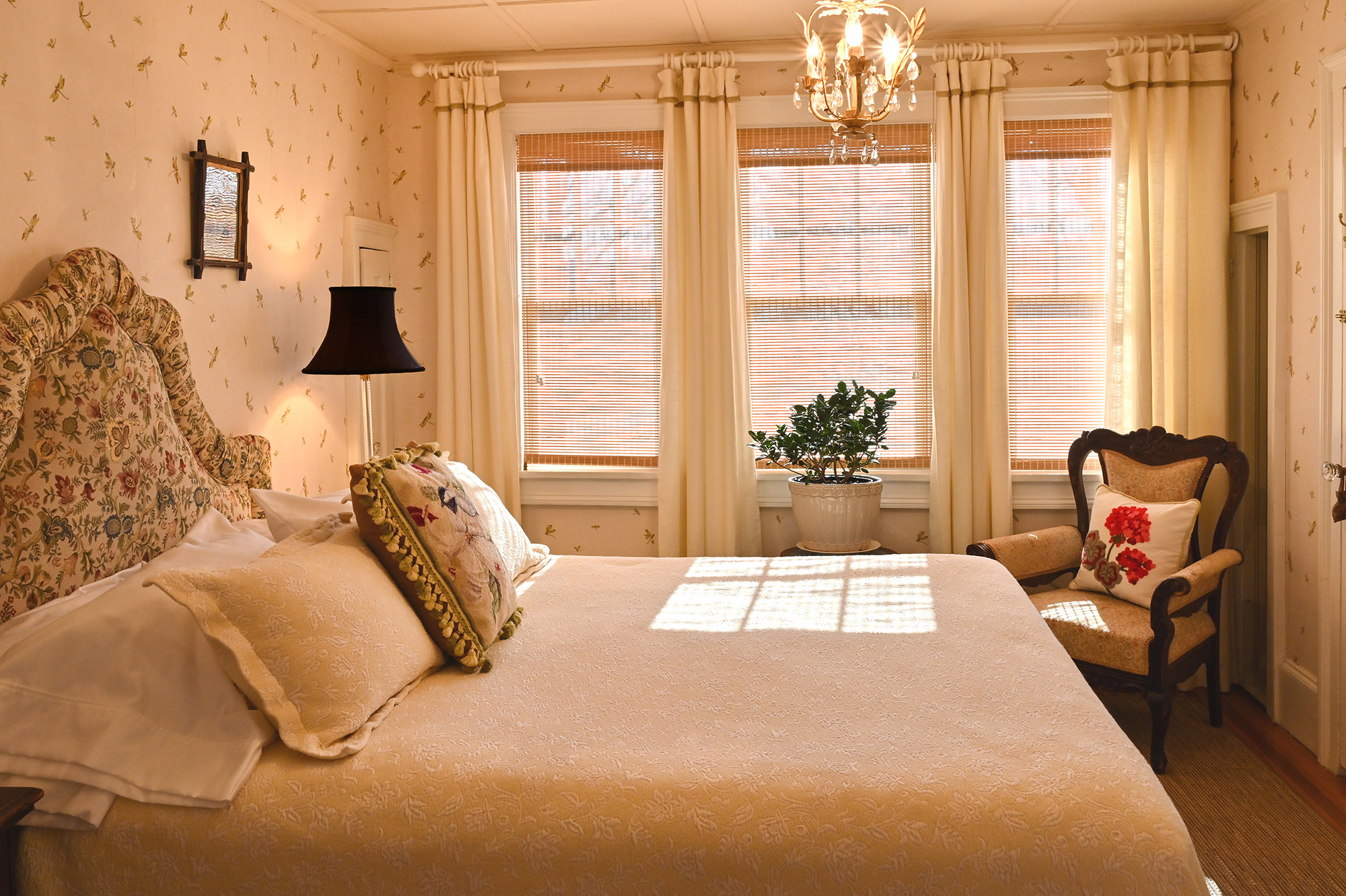 side of bed in foreground, patterned upholstered headboard. Wall of three windows, plant in front of middle window, chair with red flowered pillow in corner