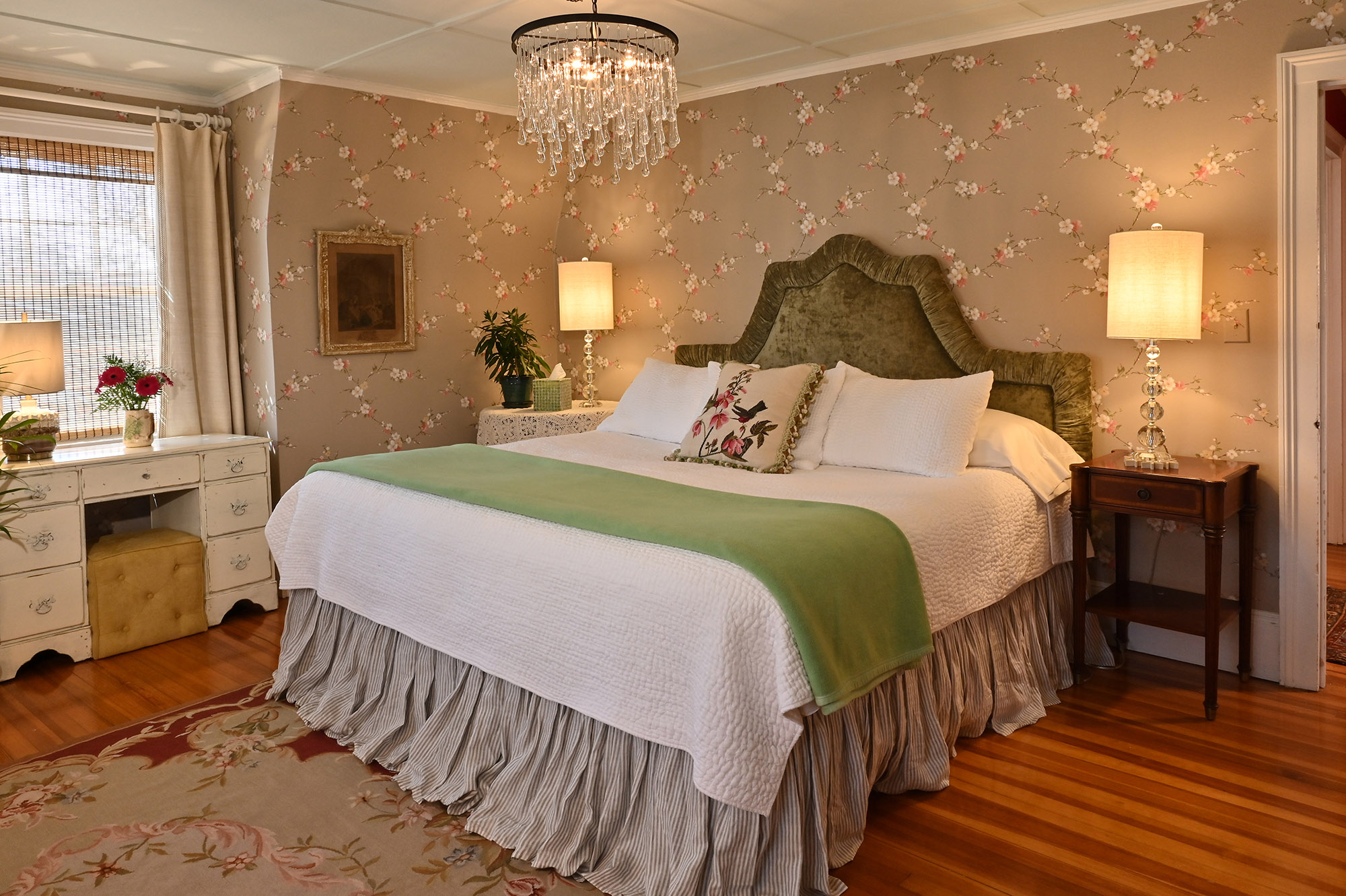 white king bed gray and white bedskirt green upholstered headboard. Antique pine wood floors. Two identical crystal lamps on each side of the bed.
