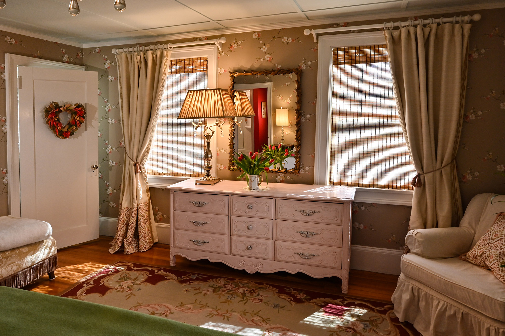 pink dresser between two windows with bamboo shades and cream colored drapery. White chair in corner taupe colored patterned rug