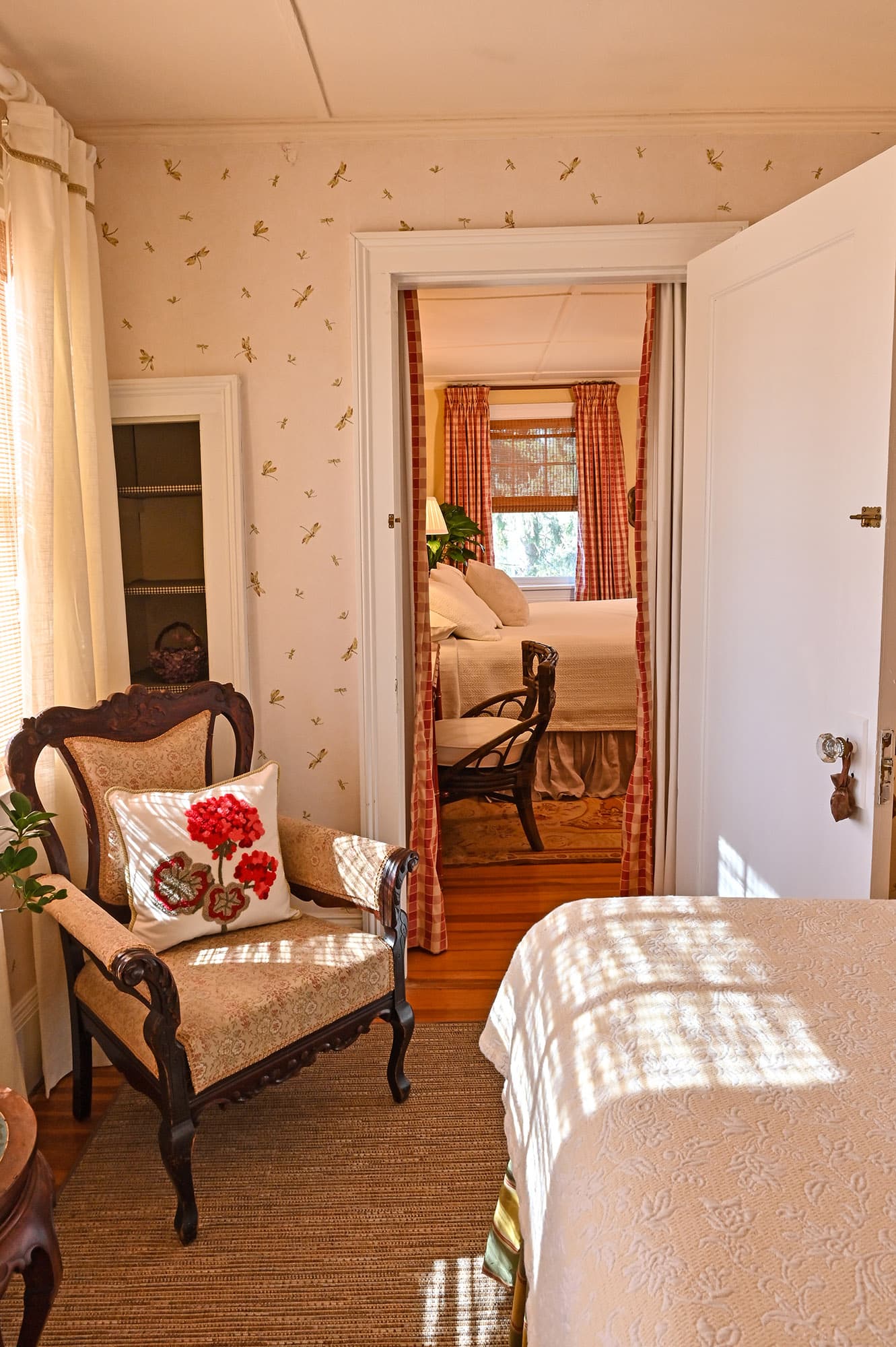 chair with red flowered pillow in corner by foot of bed. Door open leading into adjoining Sunshine Room with view of chair at desk and king bed in background in front of window