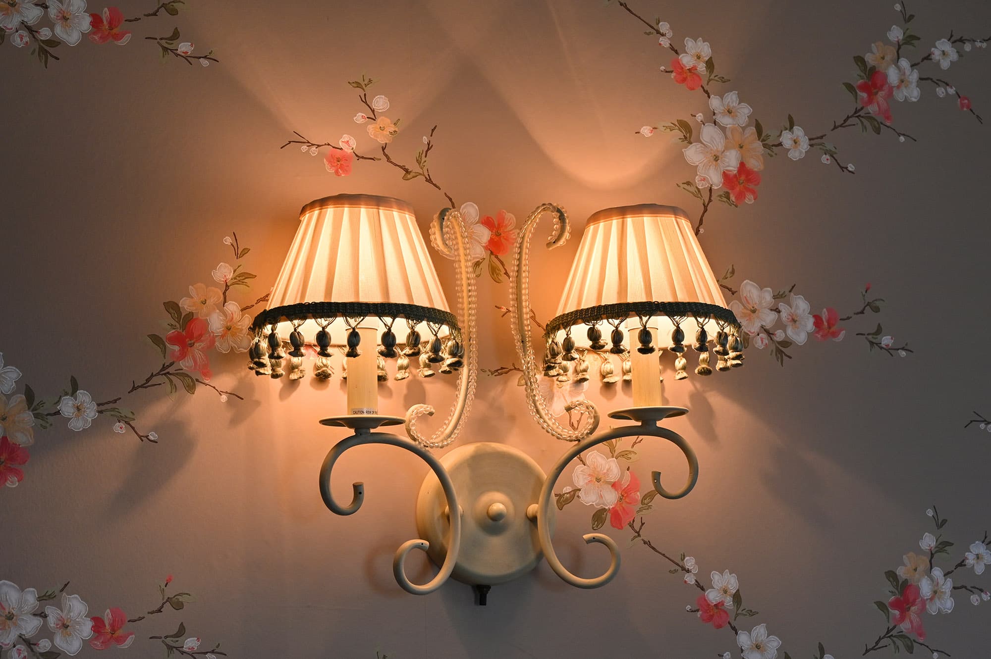 two lighted wall sconce with white shades and green tasseled bottom fringe on taupe wallpaper with pink and white vine flowers