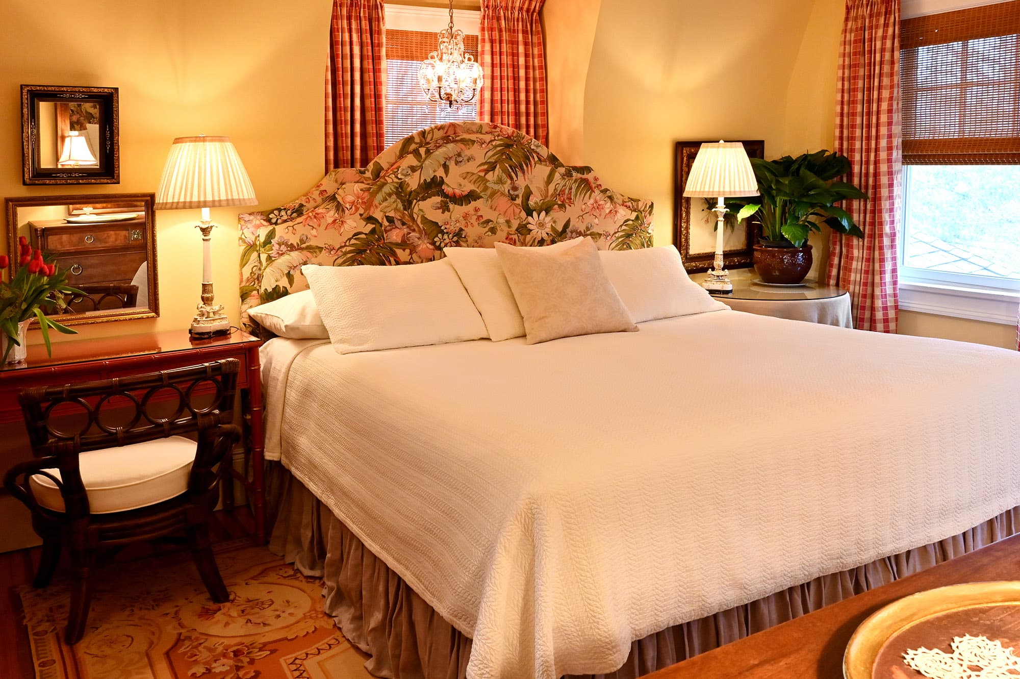 white bed with upholstered tropical patterned headboard in front of window with red curtains desk to the left round table to the right of the bed and window on the right.