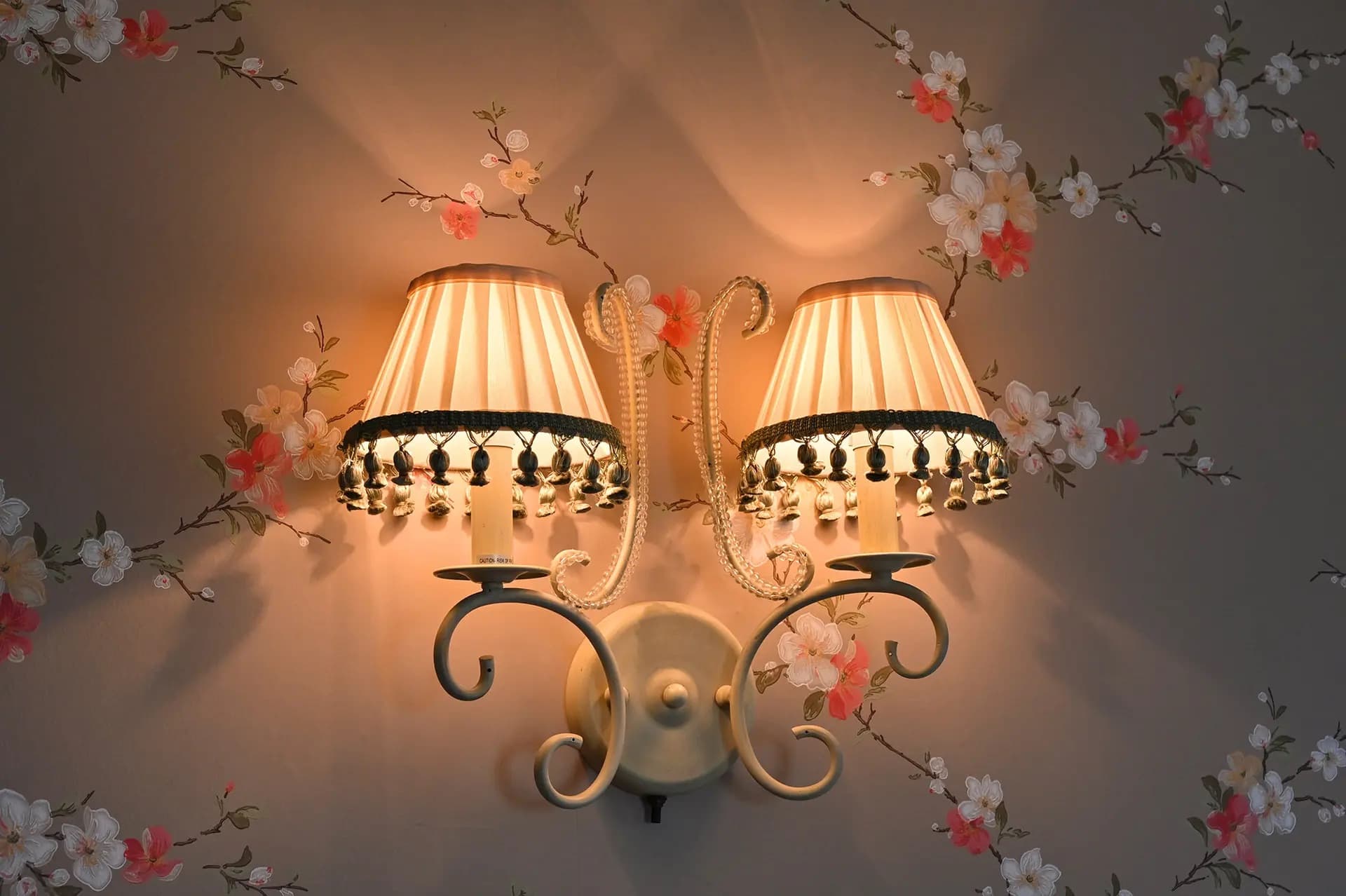 two lighted wall sconce with white shades and green tasseled bottom fringe on taupe wallpaper with pink and white vine flowers
