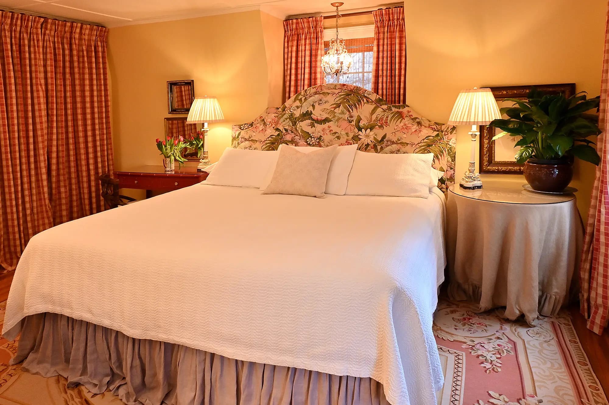 white king bed tropical patterned headboard in front of window with mauve bedskirt aubuson rug round table with tan colored tablecloth to the right of bed red desk to left of bed. Identical lamps on each side.