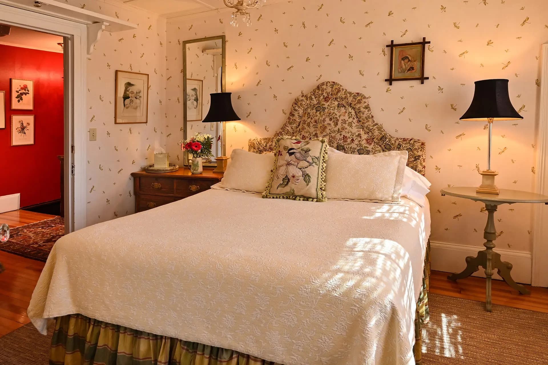 White bed green and yellow plaid bedskirt, upholstered garden patterned headboard. Dresser to left of bed mirror above. Identical lamps on each side of bed with nightstand to the right of bed