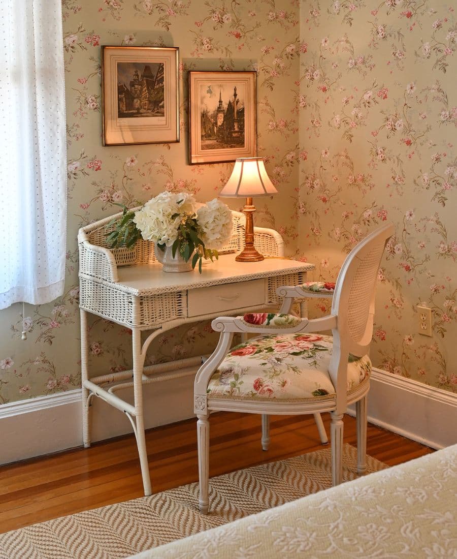A cozy corner featuring a white wicker desk with a lamp, a floral chair, and framed pictures against a floral wallpaper background.