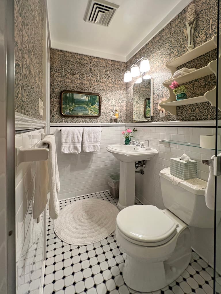 A small, stylish bathroom featuring a pedestal sink, toilet, and patterned wallpaper, complemented by decorative accents and a round rug.