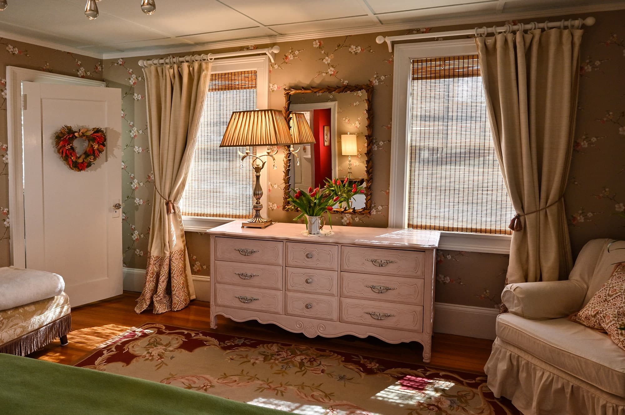 A cozy, well-decorated room featuring floral wallpaper, a dresser with a lamp and flowers, and large windows with light curtains.