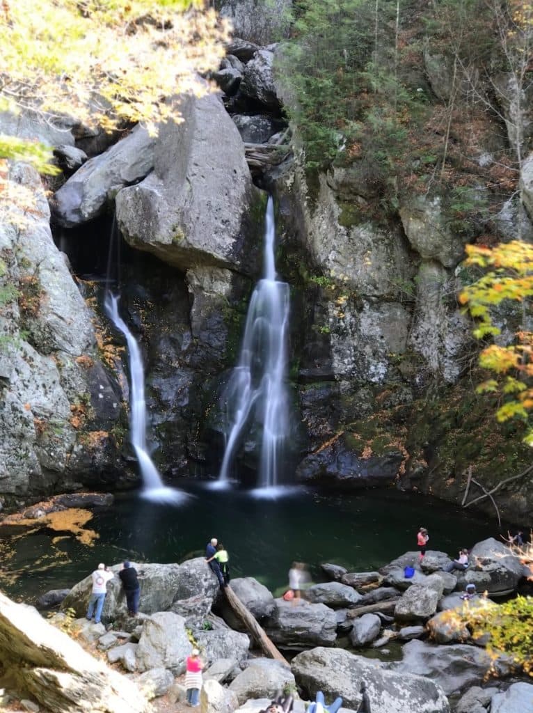 A serene waterfall cascades into a rocky pool surrounded by autumn foliage and visitors.