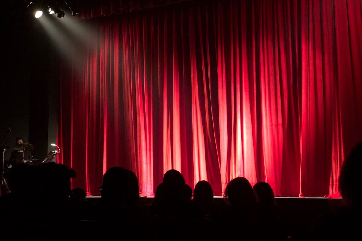 A dimly lit theater with red curtains and silhouettes of an audience in front.