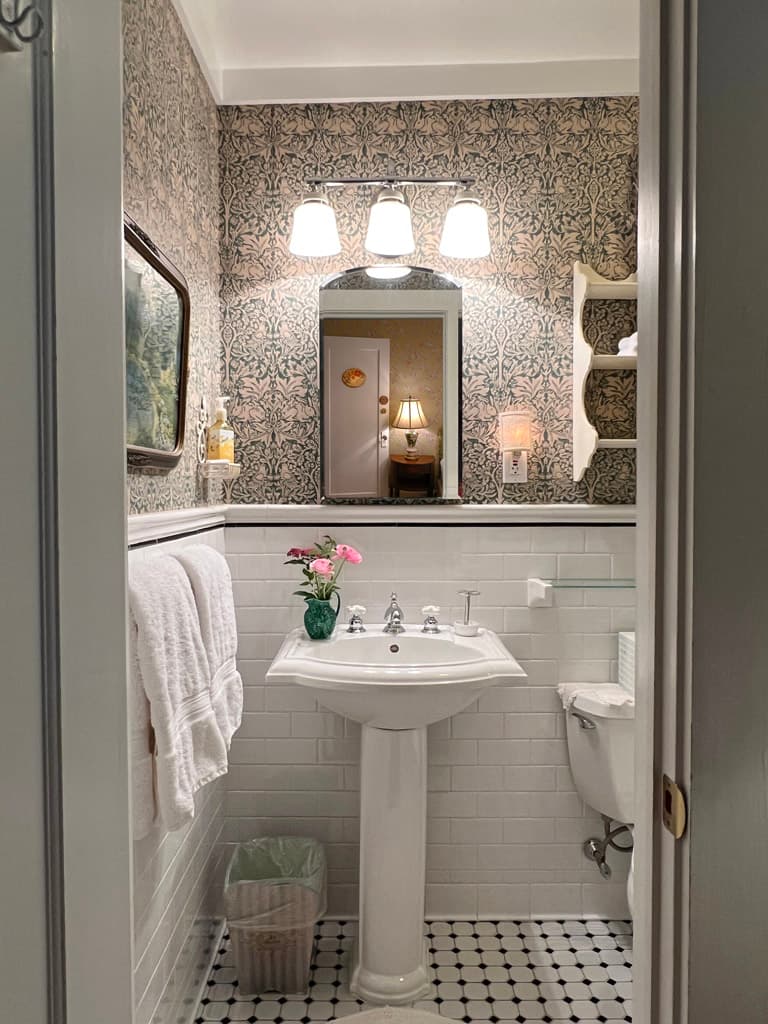 White pedestal sink white subway tile light blue wallpaper vanity lighting over mirror