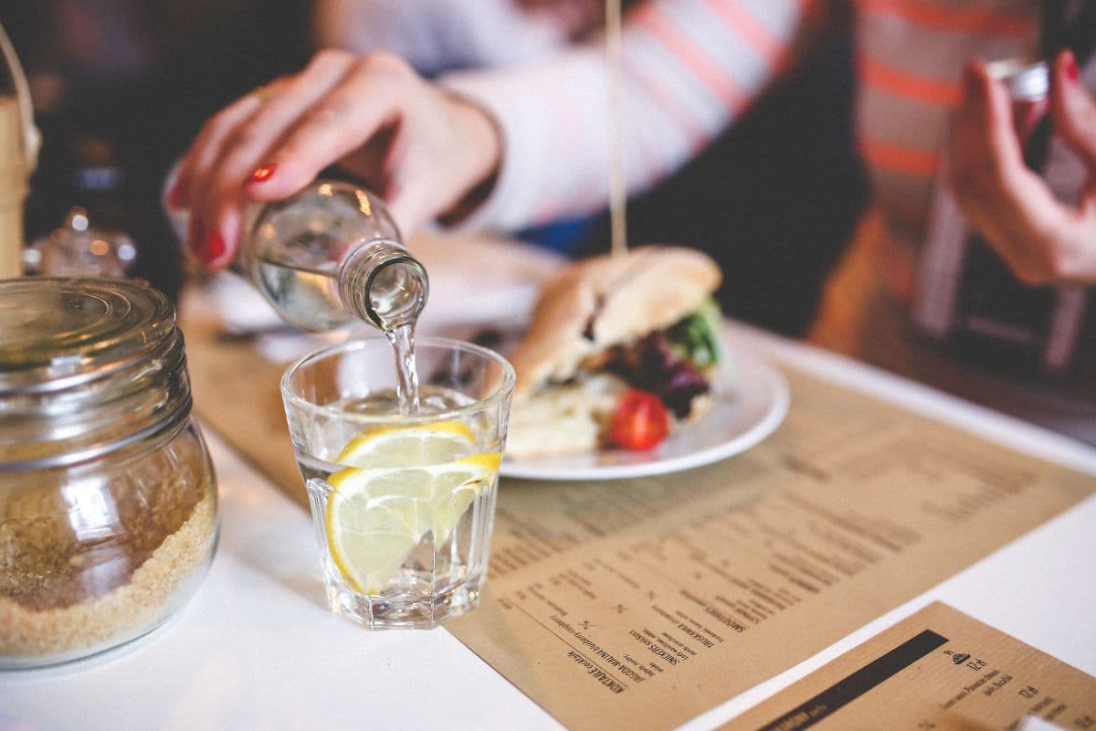 A hand pours sparkling water into a glass with a lemon slice, alongside a sandwich and a menu on a table.