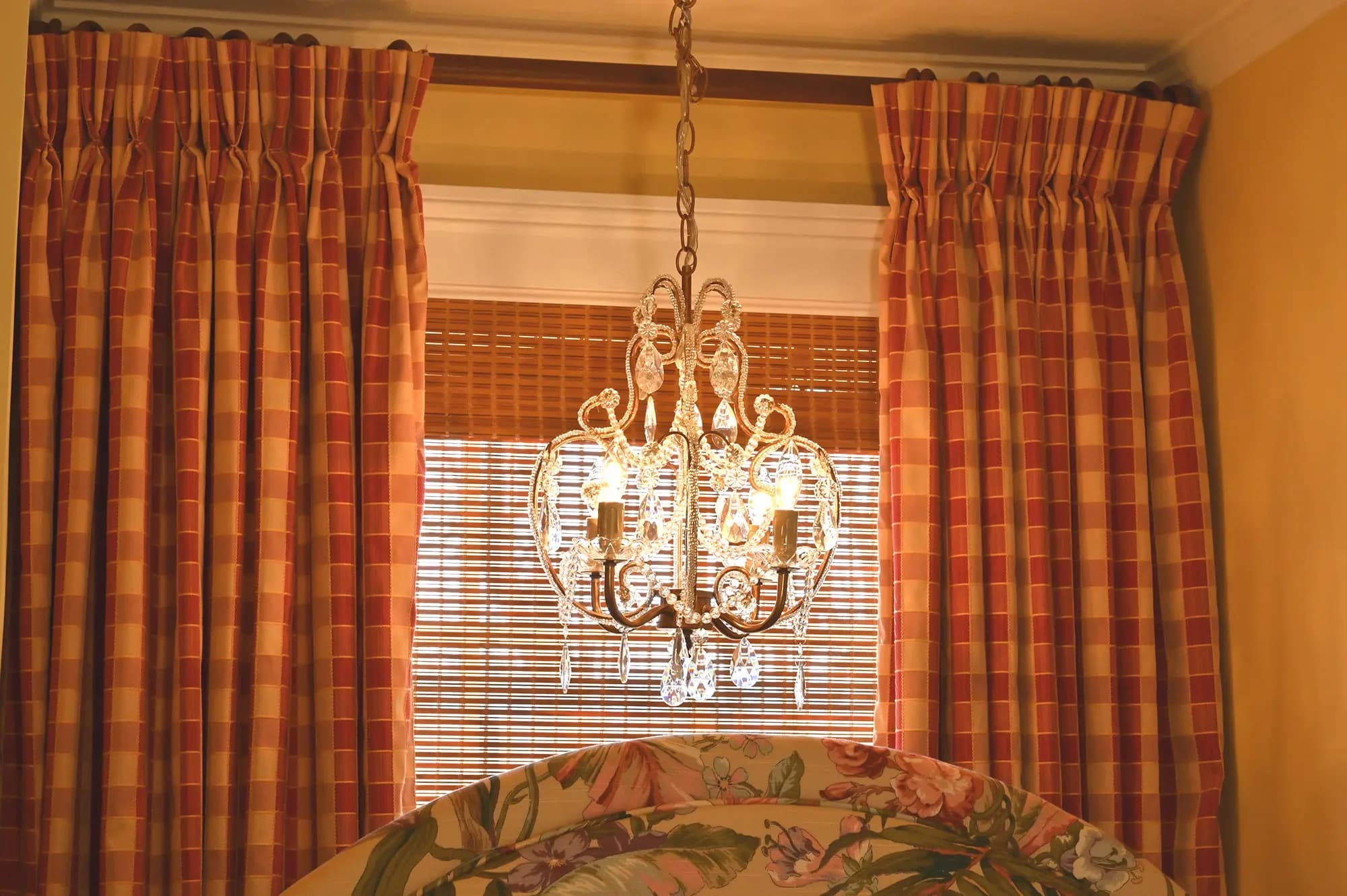 View of chandelier behind top of headboard in front of window with bamboo shade and red checked drapes