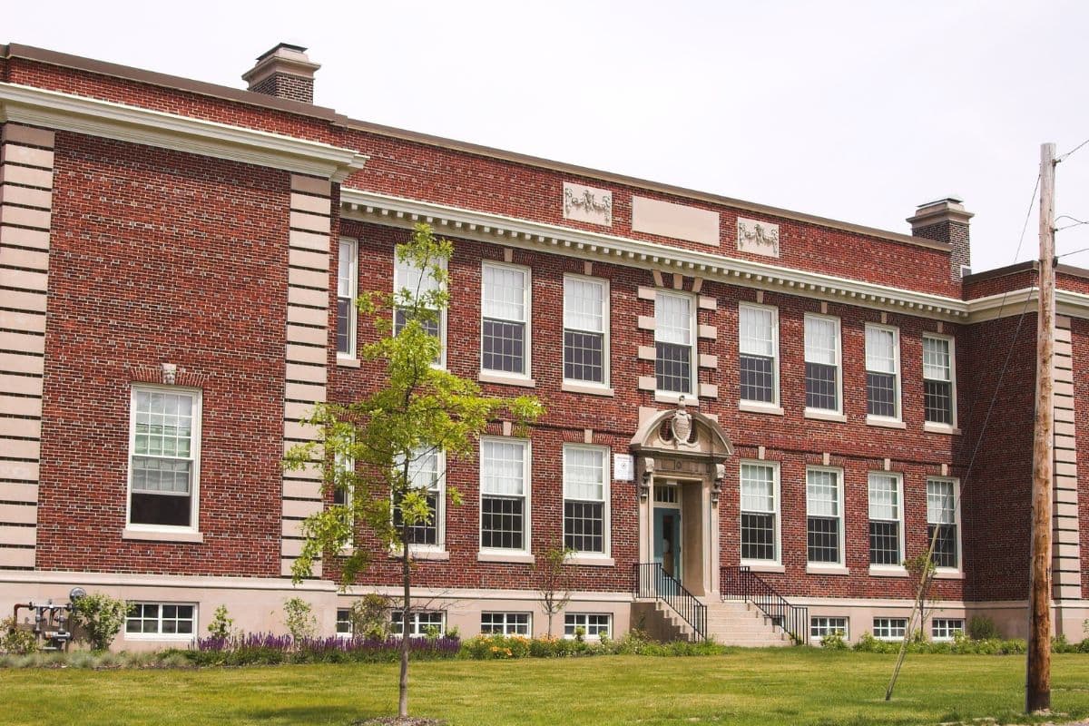 A red brick school building with large windows and a central entrance, surrounded by grass and small trees.