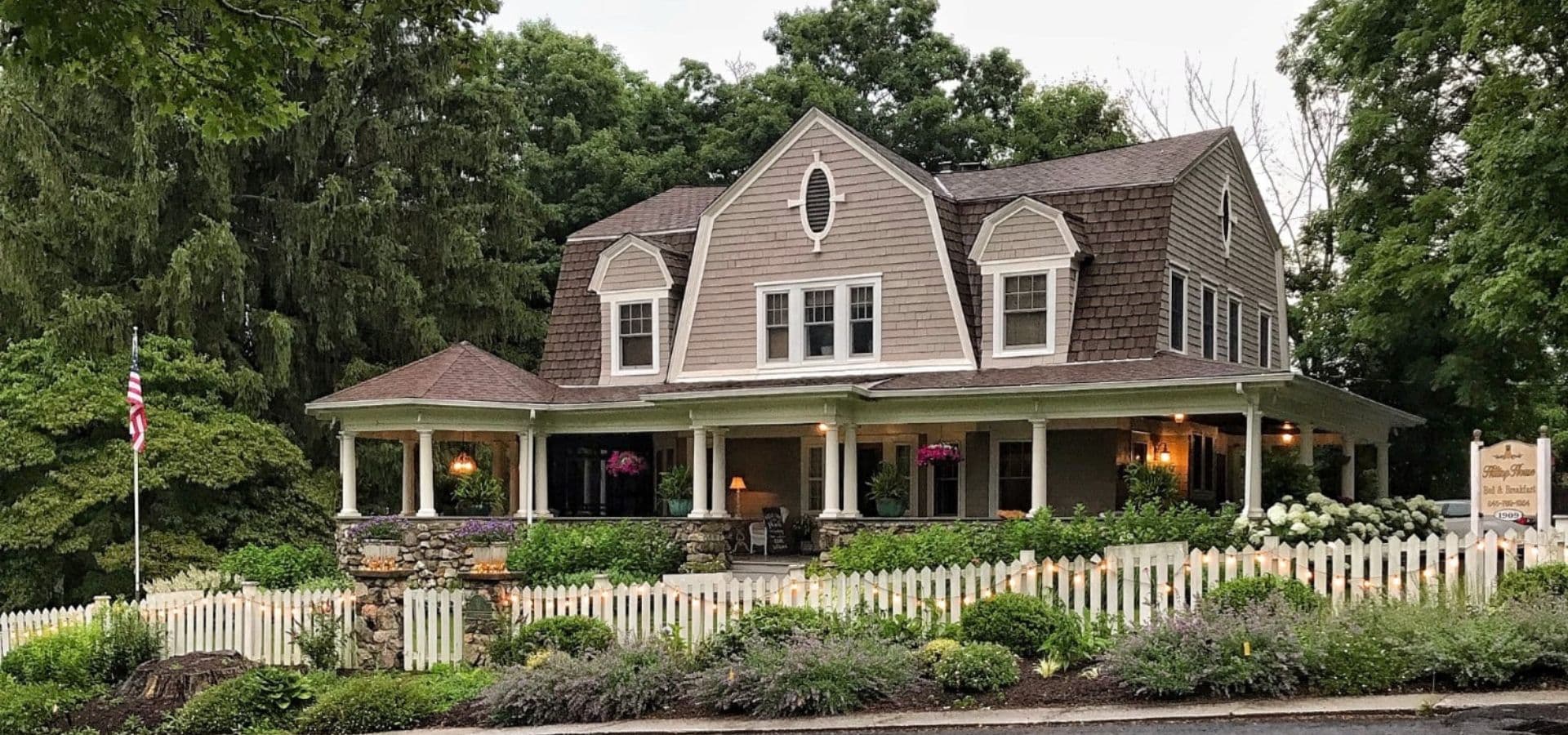 A charming gray house with a steep roof and a white picket fence surrounded by greenery.