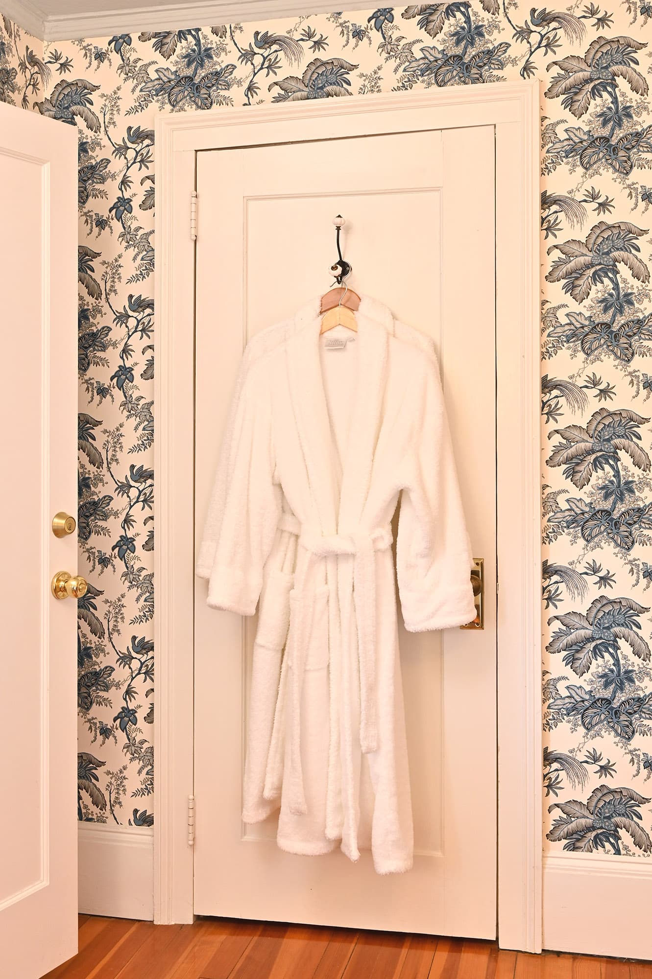 A white bathrobe hangs on a hook on a door with floral wallpaper in the background.