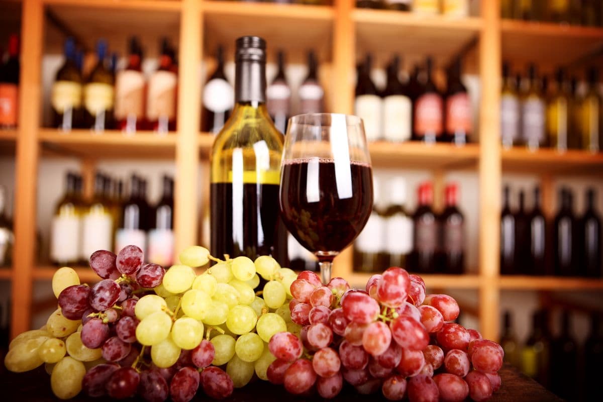 A bottle of wine and a glass of red wine beside clusters of green and red grapes, with a shelf of various wine bottles in the background.