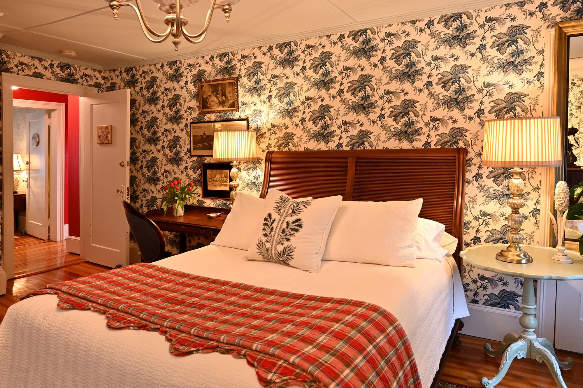 White and blue tropical themed wallpaper, queen mahogany headboard white coverlet, red and blue plaid quilt. Identical tall lamps on each side of bed desk to left and round table to right