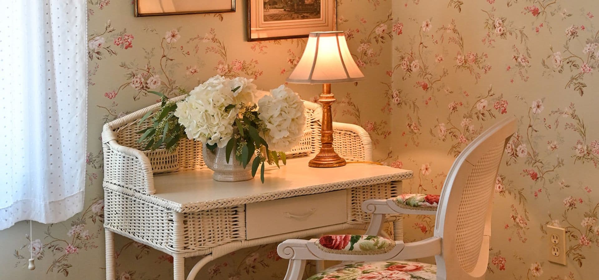 A cozy corner with a wicker desk, a lamp, and a vase of white flowers against floral wallpaper.