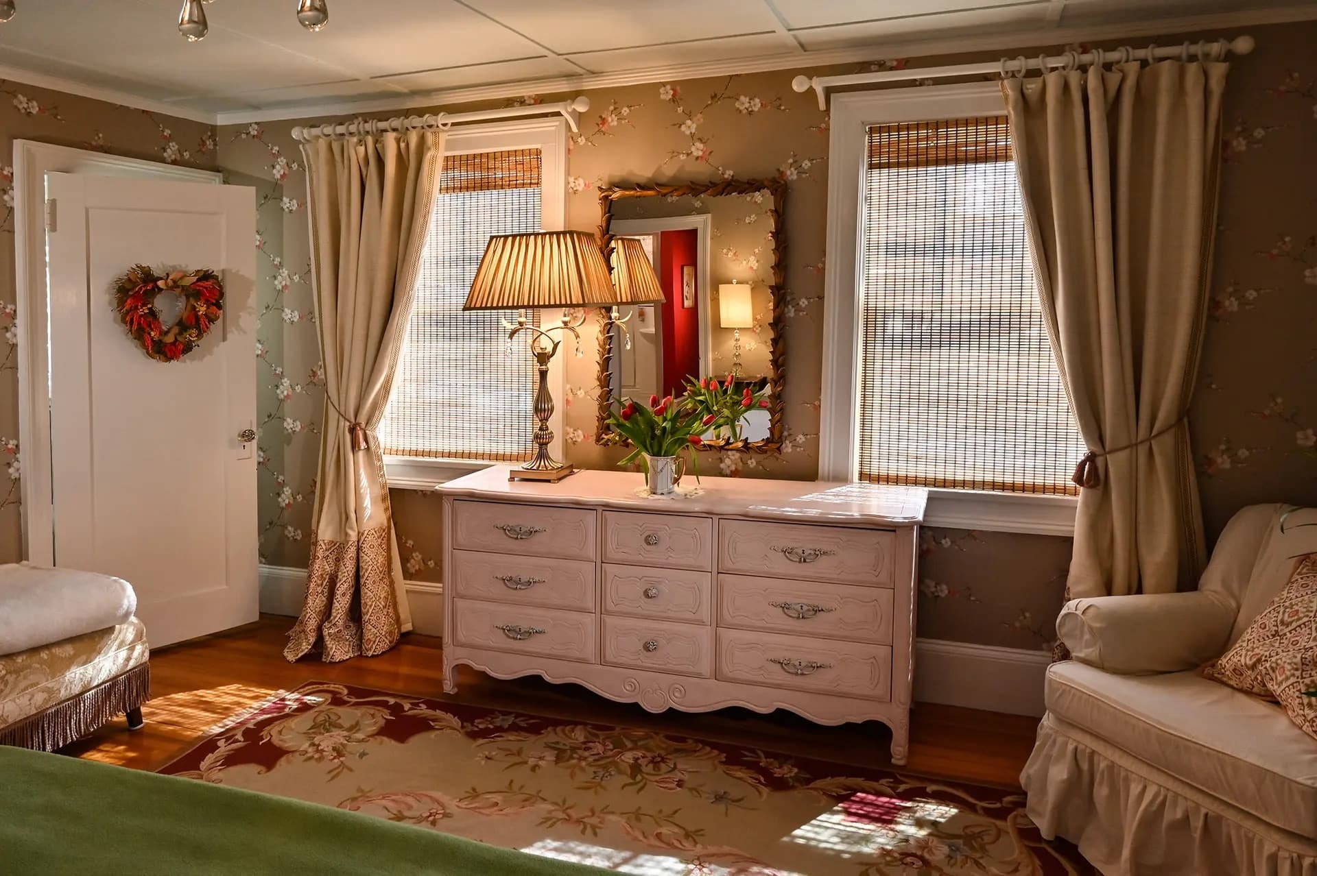 pink dresser between two windows with bamboo shades and cream colored drapery. White chair in corner taupe colored patterned rug