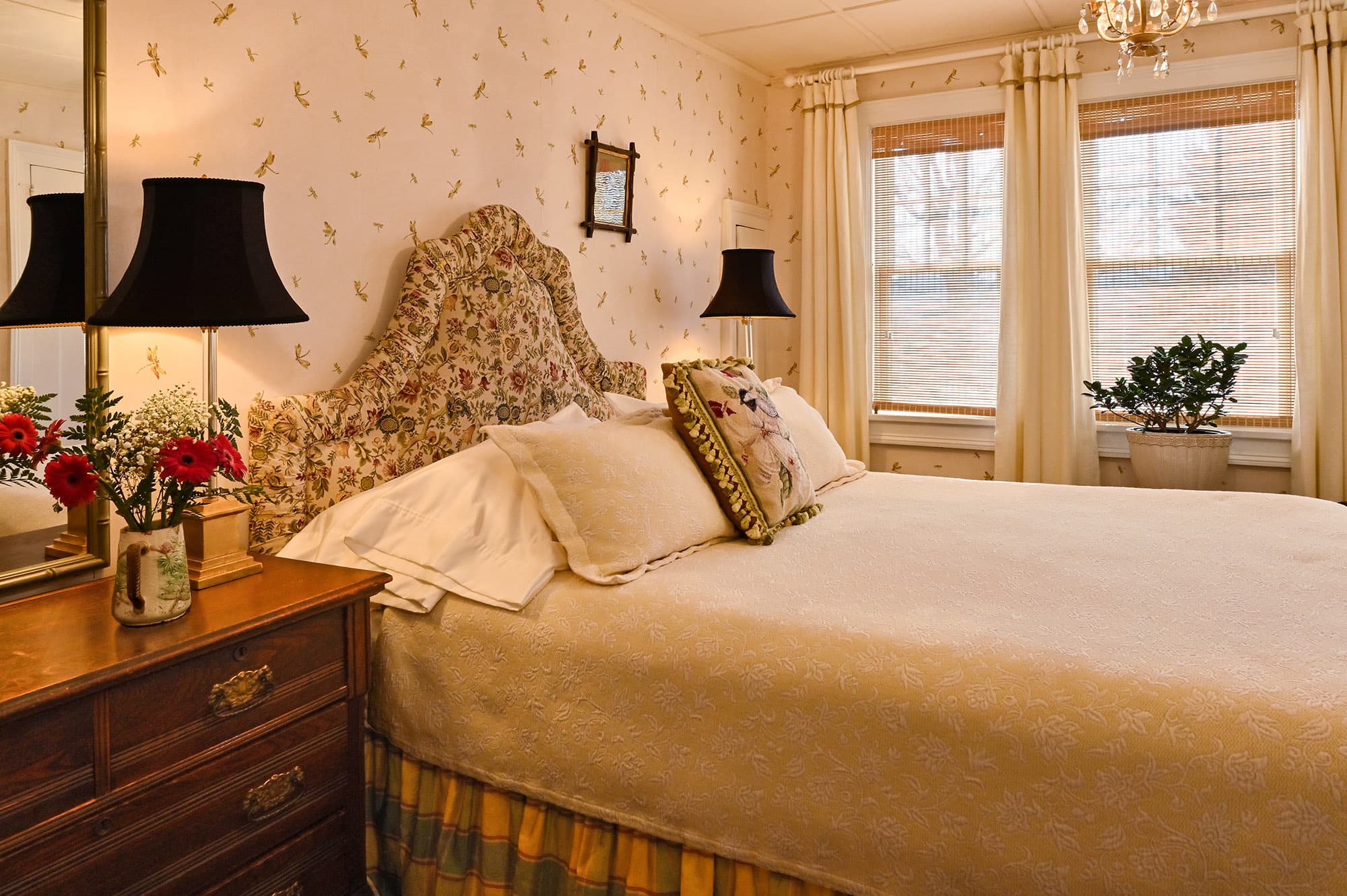 A cozy bedroom featuring floral wallpaper, a large bed with decorative pillows, and two lamps on a wooden bedside table.