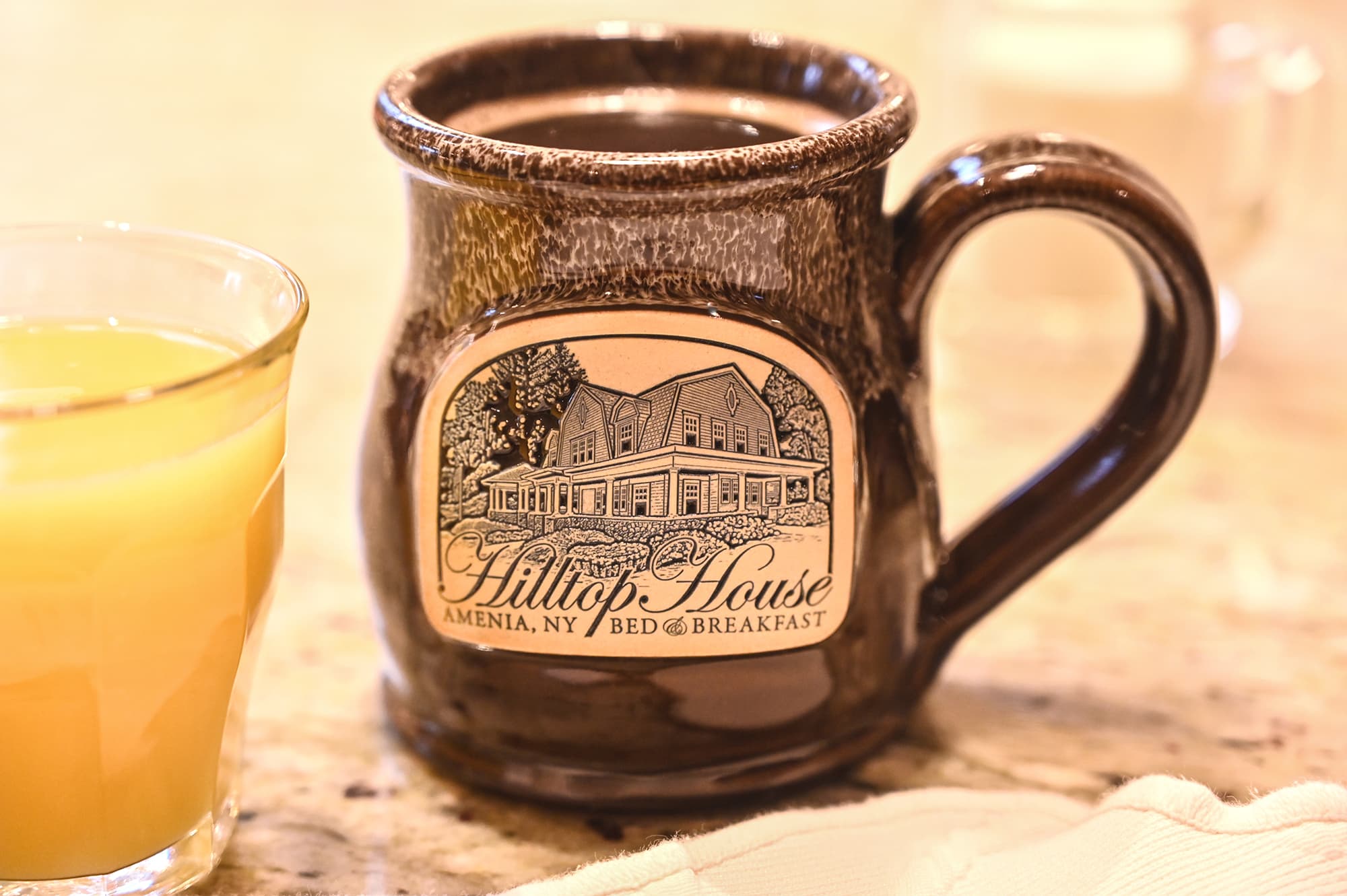 A brown ceramic mug with an illustration of Hilltop House Bed & Breakfast alongside a glass of orange juice.