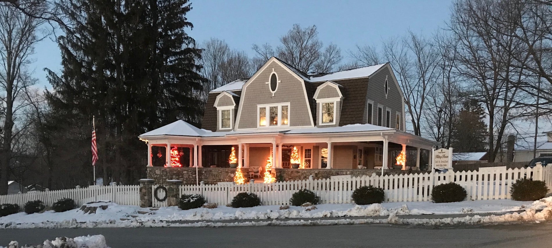 Front view in the winter of a large home with stone wrap around porch with Christmas tress, white pillars, and many windows