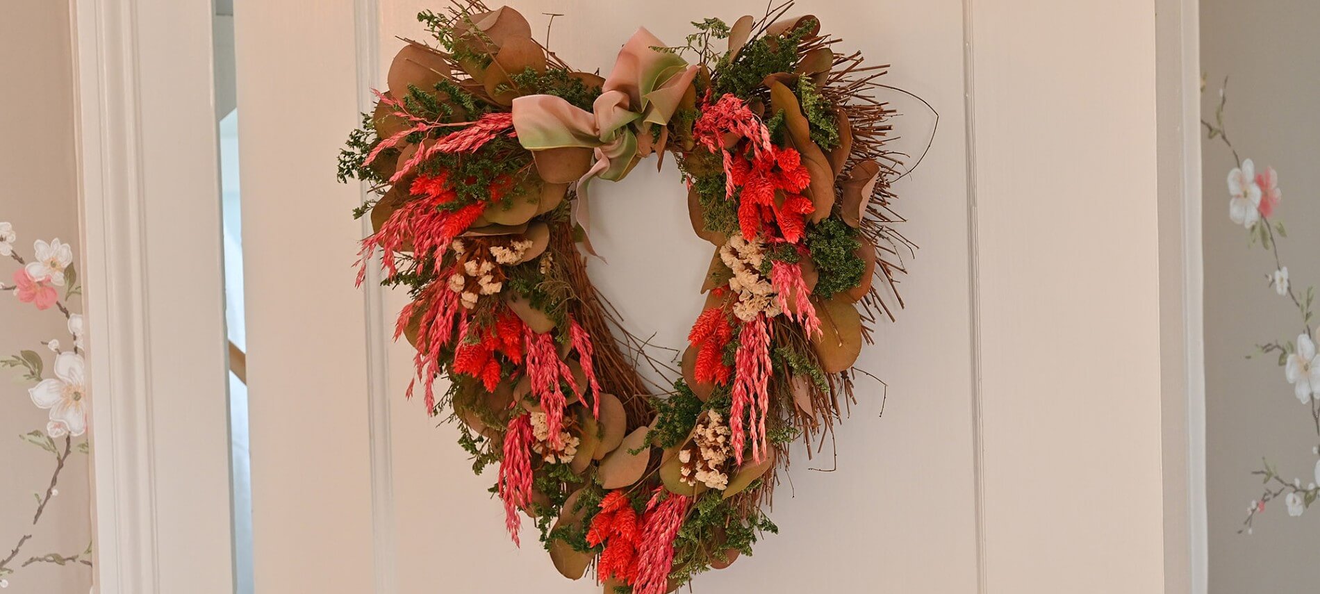 Wreath of red flowers and green leaves in the form of a heart hanging on a white door