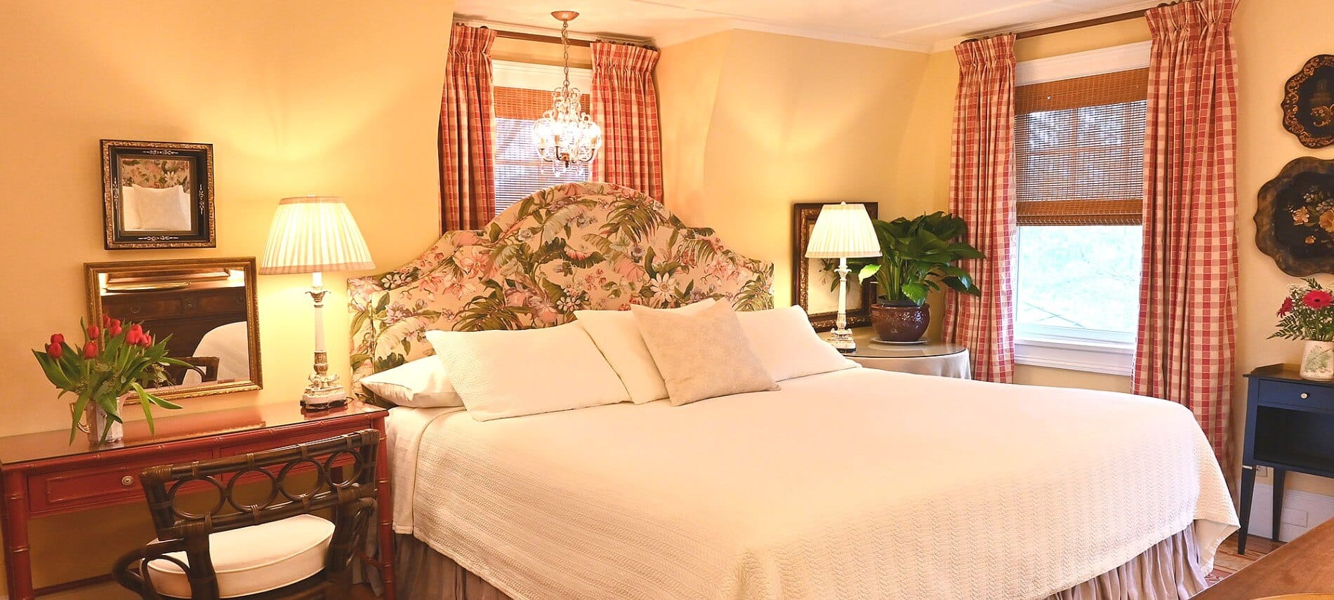 Guest room featuring a king bed with white linens, floral headboard, brown writing desk and two windows with red and white curtains