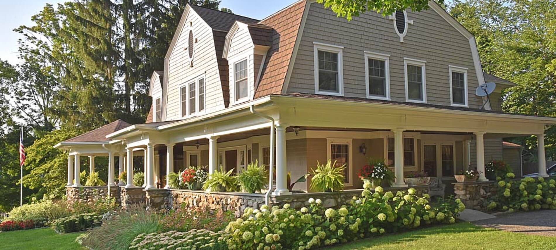 Bright sun shining on a large home with wrap around stone porch, white pillars and green landscaping