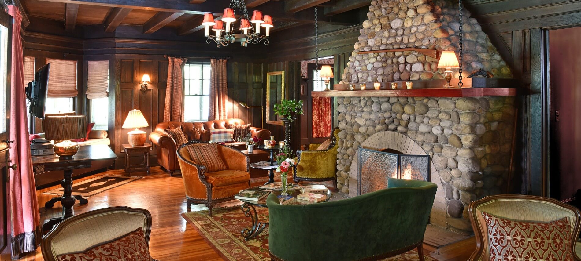 Cozy sitting room with tall rock fireplace, brown leather couch, two sitting chairs, square table and dark wood paneling on walls