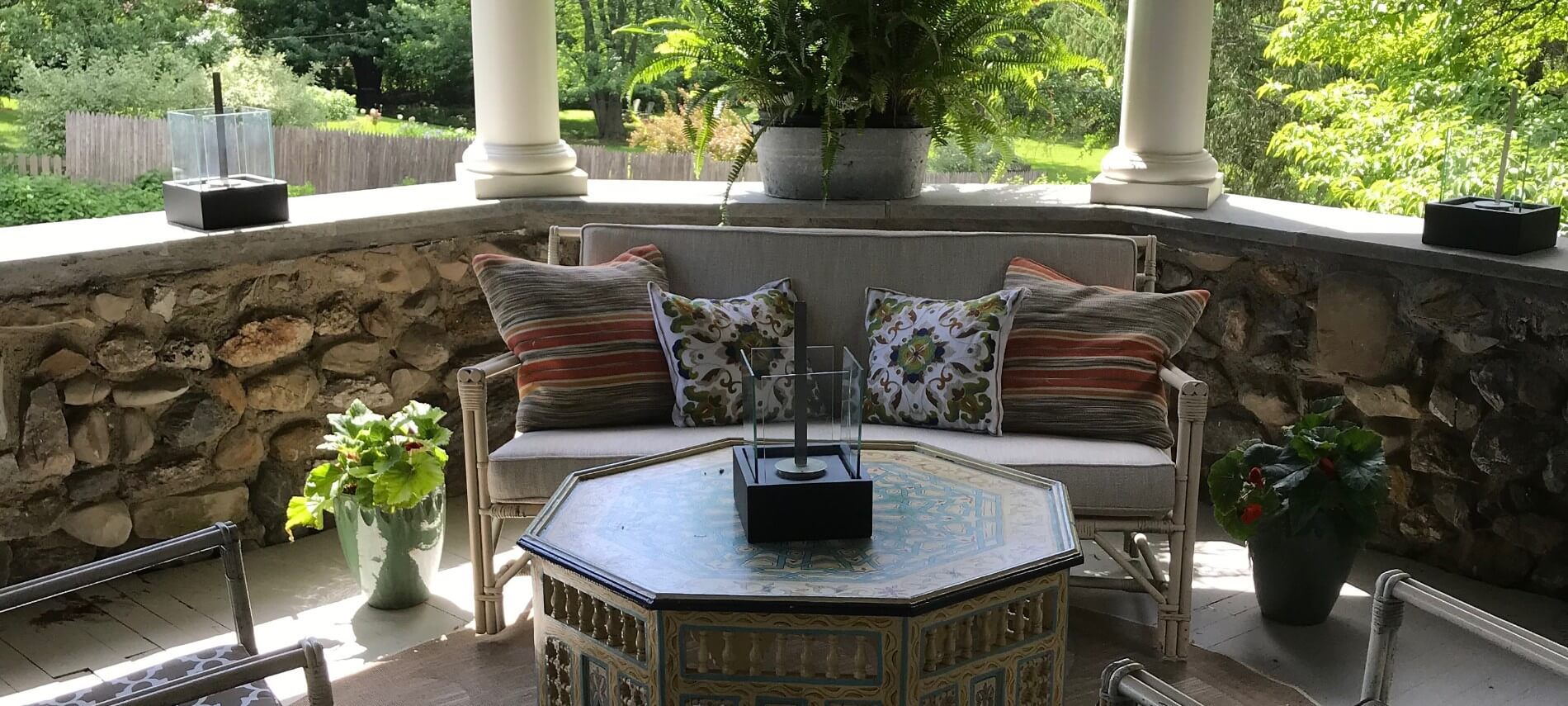 Corner of a stone porch with a wicker loveseat, octagon table and vases with green plants