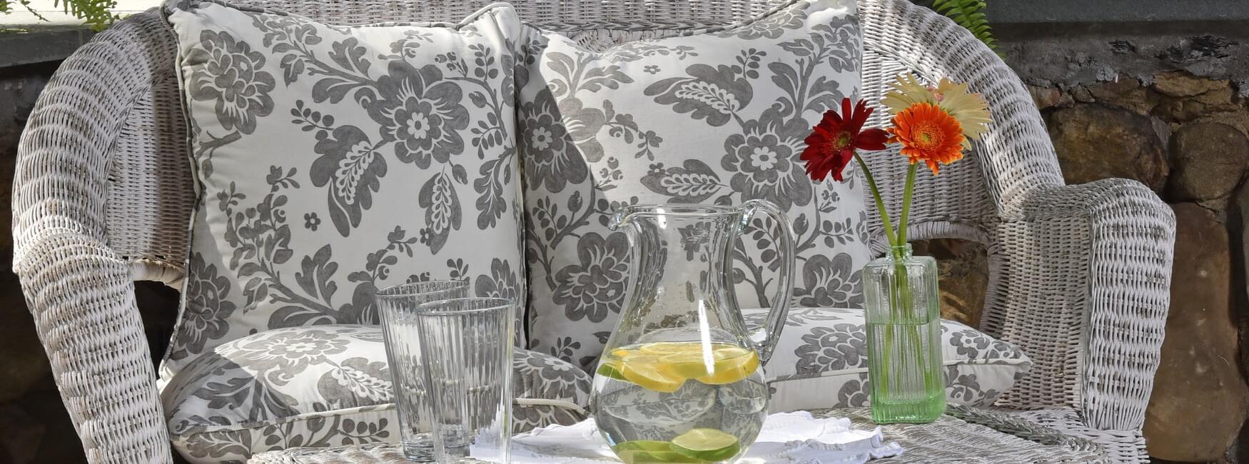 White wicker loveseat with flowered cushions with table holding pitcher of water, glasses and vase of flowers