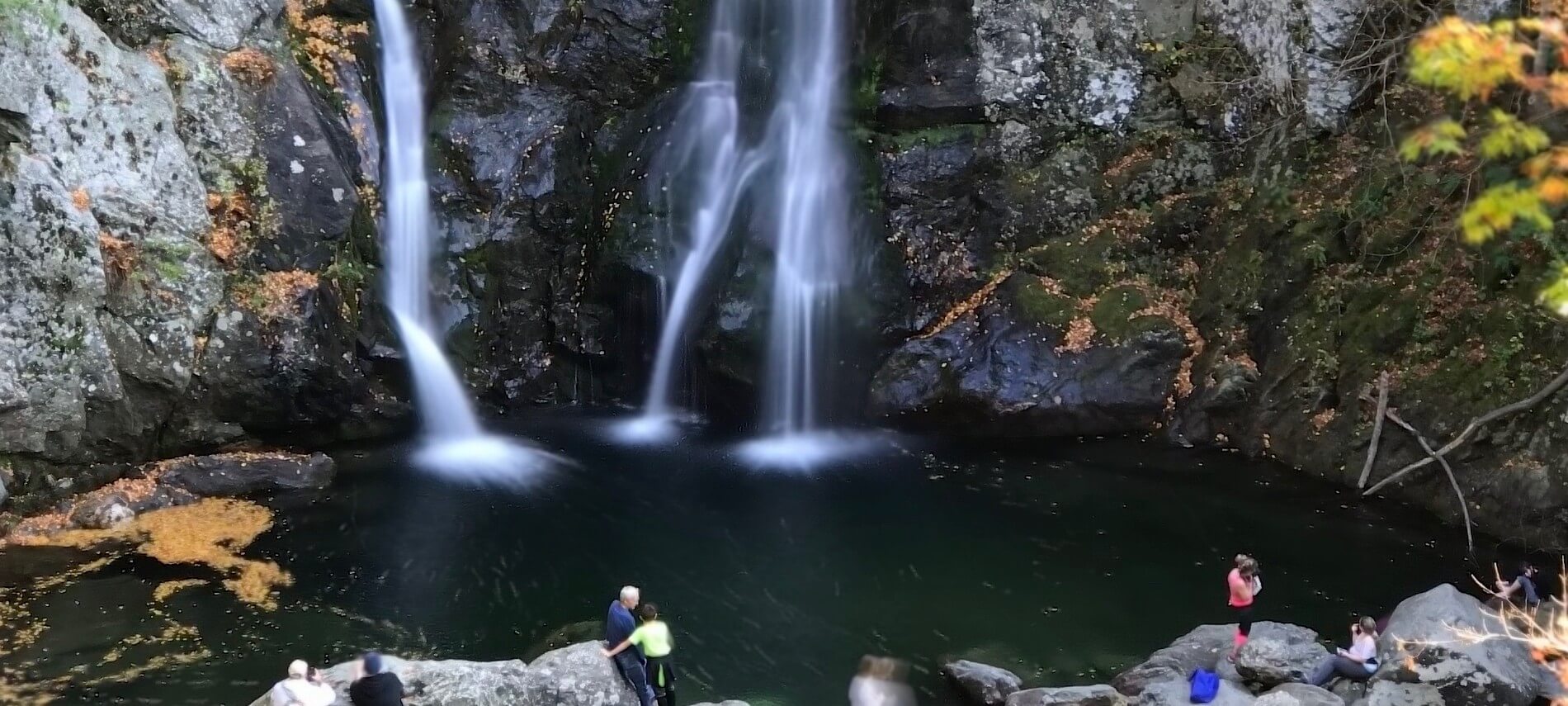 Three waterfalls cascading down into a pool surrounded by rock and trees with people standing on rocks below