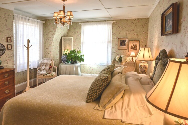 Bedroom showing queen bed with beige flowered coverlet, brown dresser, sitting chair next to round table and two bright windows
