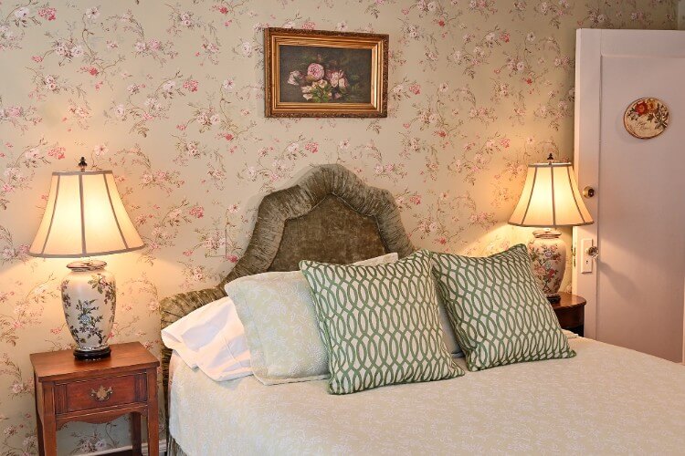 Queen bed with cream and white linens, green patterned pillows, green velvet headboard and two lamps on bedside tables