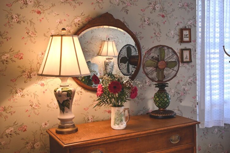 Tall brown dresser holding antique fan, tall white lamp and pitcher with red flowers against a floral wall with circle mirror