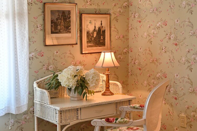 White wicker desk with white chair, one lamp and vase with two large white flowers against floral wall with two hanging pictures