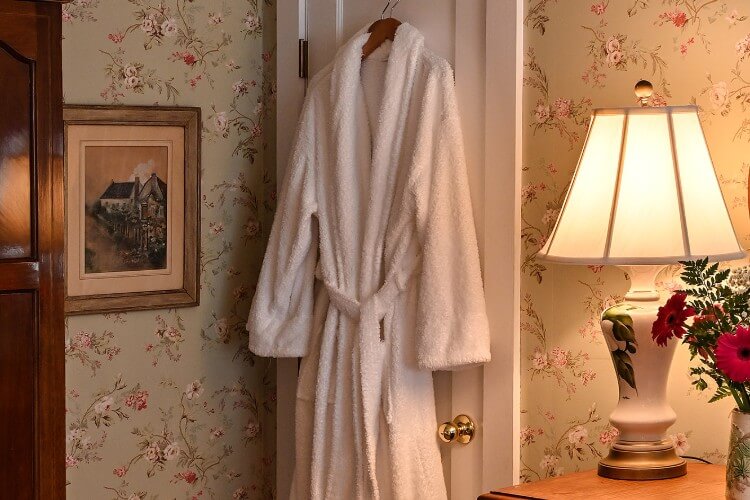 Luxurious white robe hanging on a white door in a room with floral wallpaper, and white lamp on a brown bedside table