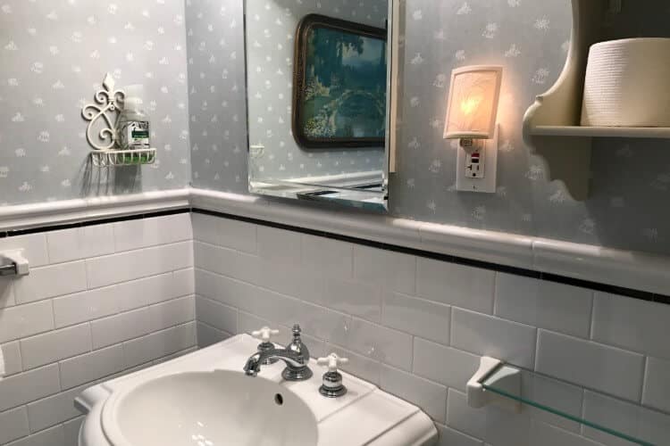 Bathroom with white tile, grey and white wallpaper, and pedestal sink with mirror and white shelving unit above