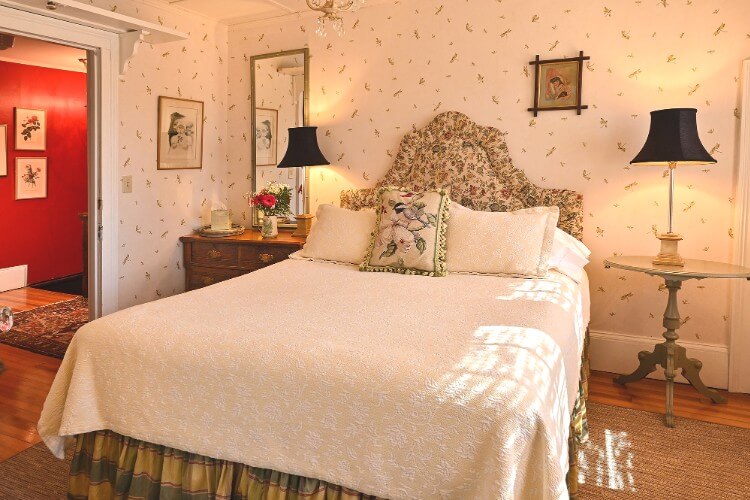 Queen bed with white linens, flowered headboard, tall black lamps and white patterned wallpaper behind