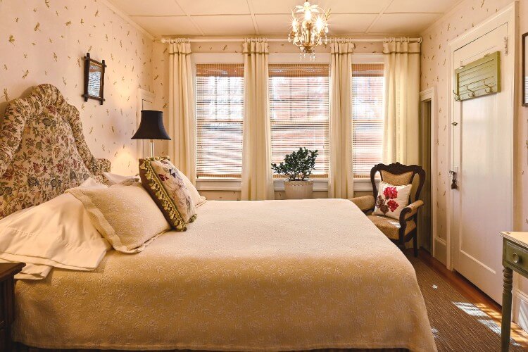 Queen bed with elegant bedding and antique sitting chair in front of three tall windows with white curtains