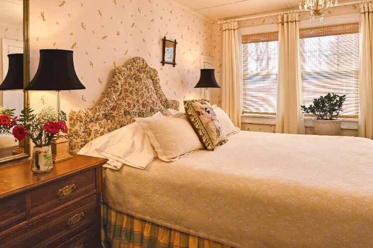 Queen bed with flowered headboard, brown dresser with tall black lamp next to bright windows with white curtains