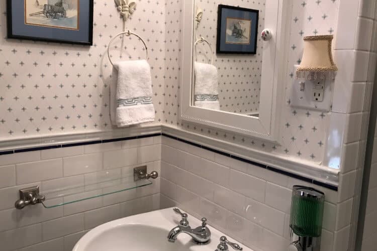 Bathroom with white tile, pedestal sink with white mirror above and blue and white wallpapered walls