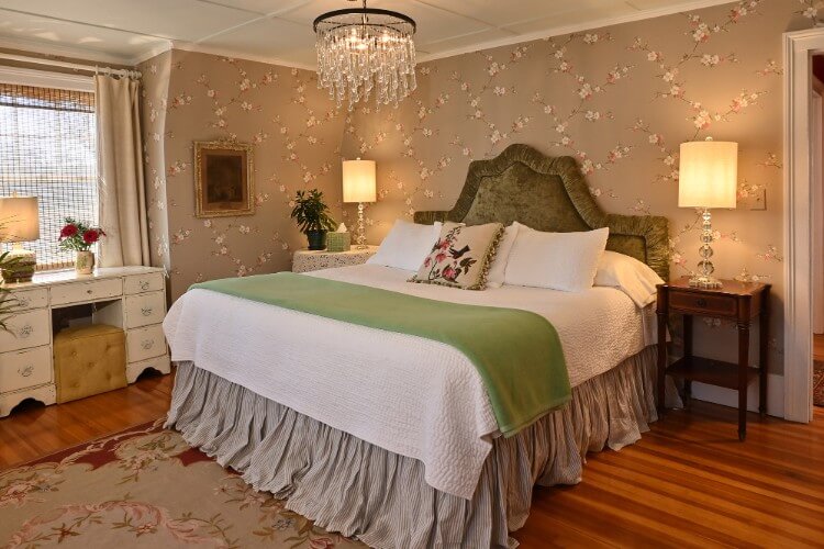 King bed with white and green bedding in elegant room with chandelier light and brown flowered wallpaper