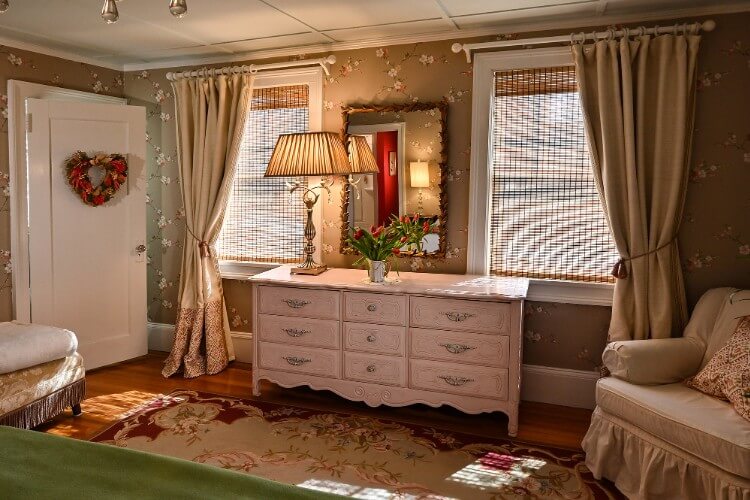 White dresser with mirror in a guest room with upholstered sitting chair and two large windows with beige curtains