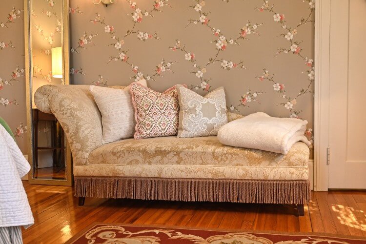 Chaise lounge with gold patterned upholstery, three pillows and a white folded blanket against flowered wallpaper wall