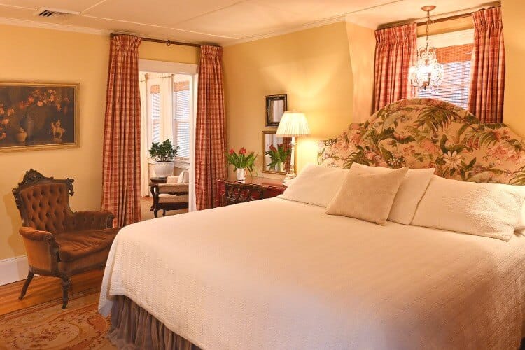 Peaceful guest room with antique furniture, large bed with white linens and floral headboard, and doorway into a sitting room