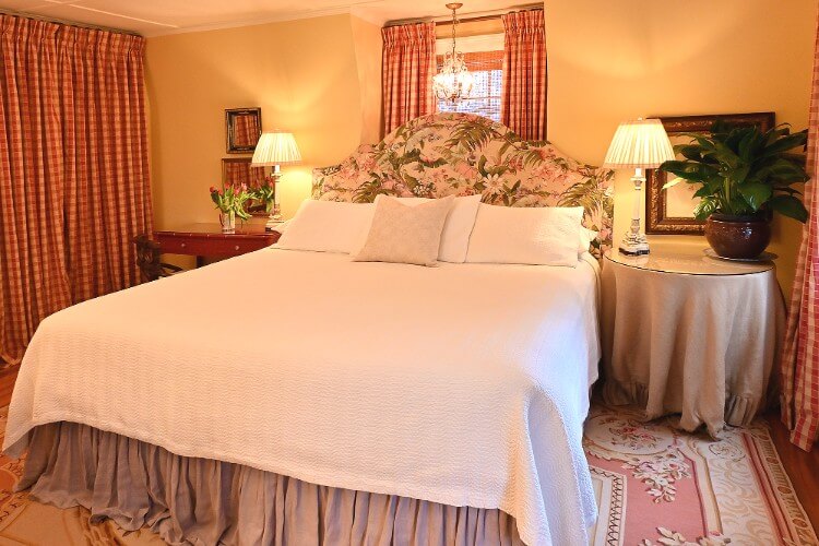 Elegant king bed with white comforter, floral headboard, and two softly lit lamps by window with red and white curtains